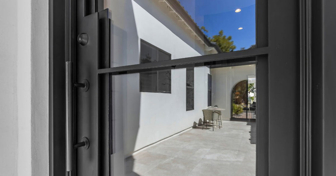 Black metal and glass door frames a sunlit courtyard with white walls, tiled flooring, and a small table and chair.