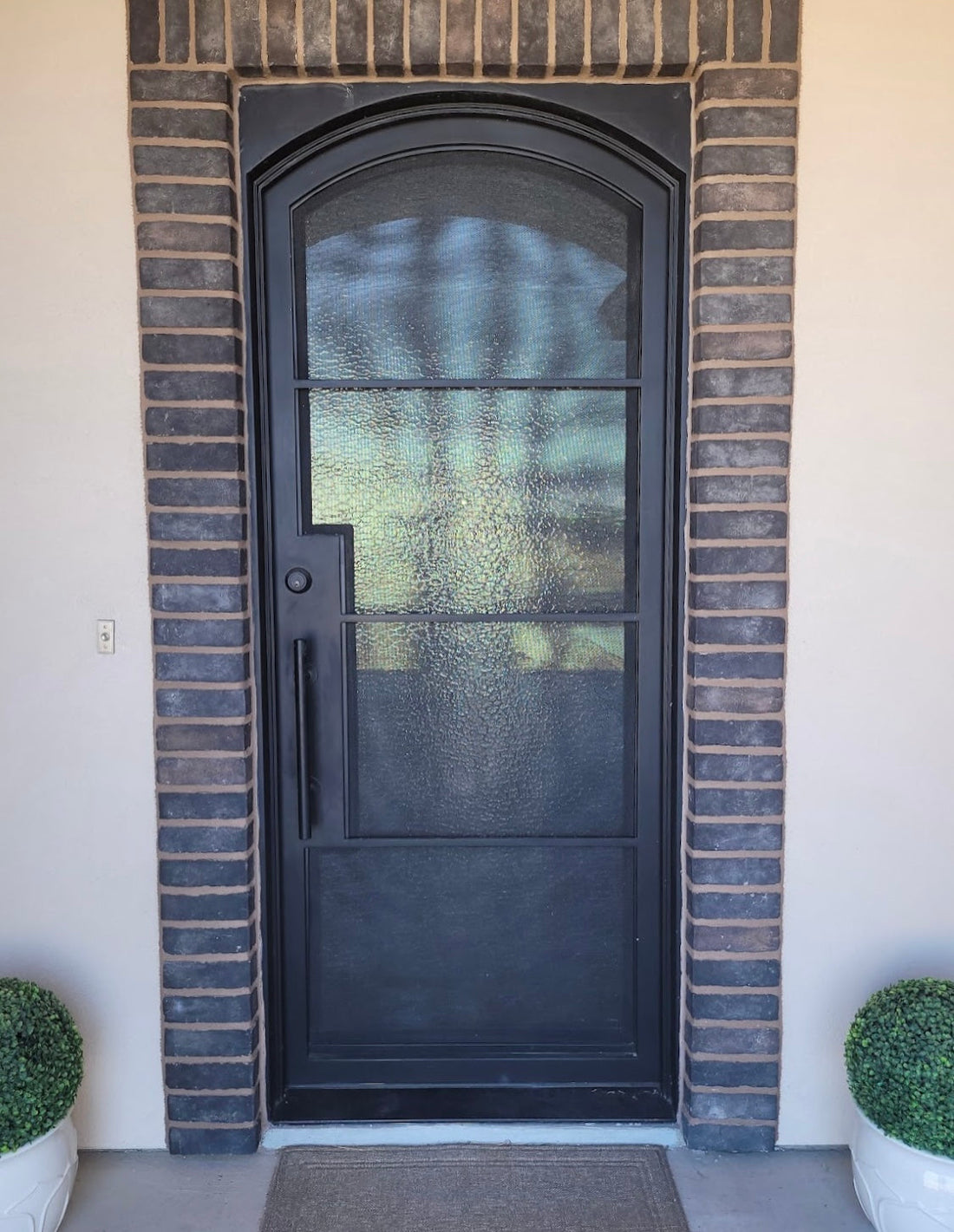 Black arched front door with textured glass, framed by dark brick and flanked by matching potted shrubs.