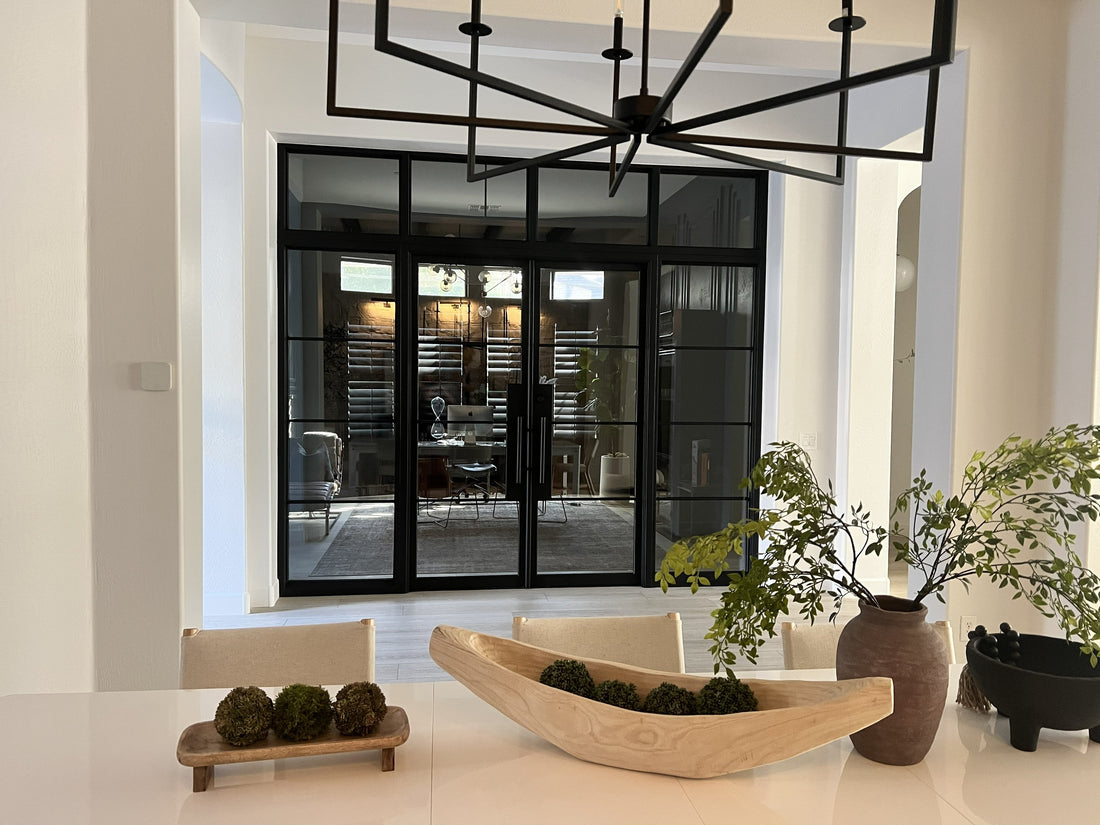 Black-framed glass doors and transom panels enclose a home office viewed from a bright, modern dining room.