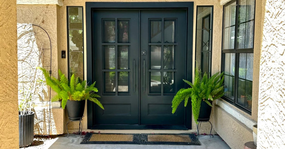 A black double front door with glass panels, two doormats, potted ferns, and windows beside a beige porch.