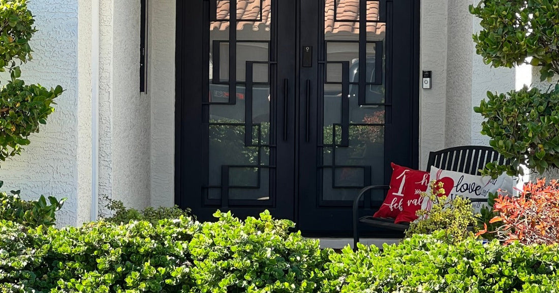 A front porch with black double glass doors, green shrubs, potted trees, and a bench with red pillows.