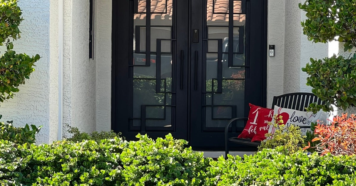 A front porch with black double glass doors, green shrubs, potted trees, and a bench with red pillows.